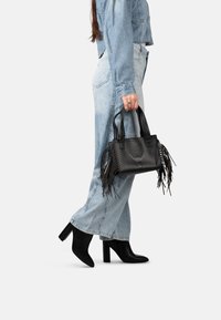 Borsa in pelle nera con accenti di frange, manici intrecciati e logo impresso. Abbinata a una giacca di jeans azzurro chiaro e jeans a gamba larga.