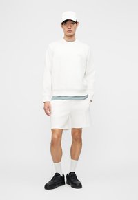 Νεαρός άνδρας με λευκό φούτερ, λευκά σορτς, λευκό καπέλο, λευκές κάλτσες και μαύρα αθλητικά παπούτσια, που στέκεται μπροστά σε μονόχρωμο λευκό φόντο.