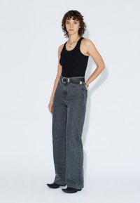 Top nero senza maniche, jeans in denim grigio a vita alta, a gamba larga, con passanti per cintura e una cintura nera. Modella in posa su uno sfondo bianco.