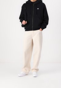 Felpa nera con zip e tasca frontale, pantaloni a gamba larga color crema e sneakers bianche. Presenta un piccolo logo bianco sul petto.