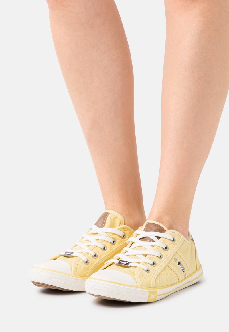 Yellow canvas sneakers with white laces, rubber toe caps, and a brown logo patch. Features a low-profile design and textured fabric.