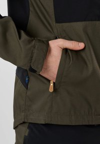 Main dans la poche avant d'une veste verte et noire avec une petite étiquette drapeau bleu et jaune sur le côté.