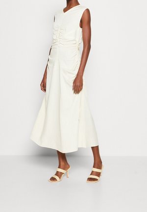 Kobieta nosząca bezrękawową, kościelnobiałą sukienkę midi z marszczonymi detalami, zestawioną z beżowymi sandałami na obcasie, stojąca na białym tle.