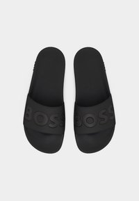 Ζεύγος μαύρων σανδαλιών με υφή στα  πέλματα και φαρδιές λουρίδες με χαραγμένα γράμματα "BOSS" στην κορυφή.