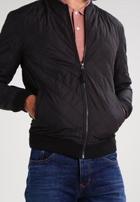 Schwarze gesteppte Bomberjacke mit einem Reißverschluss, gerippten Bündchen und Saum, getragen über einem rosa Poloshirt, kombiniert mit blauen Jeans.