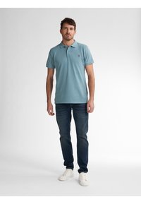 Camisa polo azul claro de manga corta con una tapeta de botones, combinada con unos vaqueros oscuros de mezclilla y zapatillas blancas, de pie contra un fondo neutro.