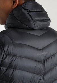  Černá péřová bunda s vysokou prošívanou kapucí. Obsahuje šicí švy ve tvaru V a logo "Mammut" tištěné na zadní straně. Hladký, lesklý materiál.