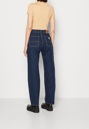 Person trägt beige kurzärmeliges Oberteil, dunkelblaue hoch taillierte Jeans mit weitem Bein und großen Gesäßtaschen sowie schwarze Plateauschuhe, Schau in die andere Richtung.