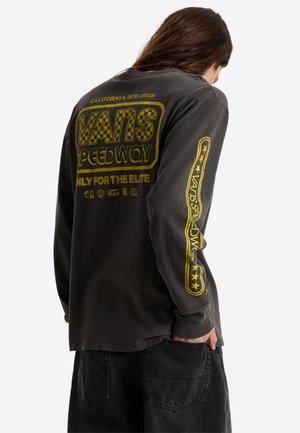 Persona con pelo largo que lleva una camiseta oscura de manga larga Vans Speedway con texto y gráficos amarillos de cuadros en la espalda y la manga.