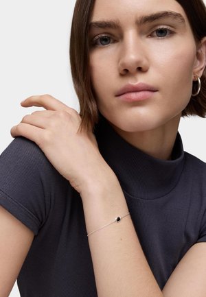 Pulsera de plata de ley con un pequeño acento de piedra negra. Modelo vistiendo un top de cuello alto oscuro con canalé. Textura de piel suave visible.