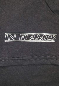 Czarny t-shirt z tkaniny, na którym znajduje się biały haftowany napis "IN FLAMES" w pogrubionej, bezszeryfowej czcionce, z widocznym detalem szwów.
