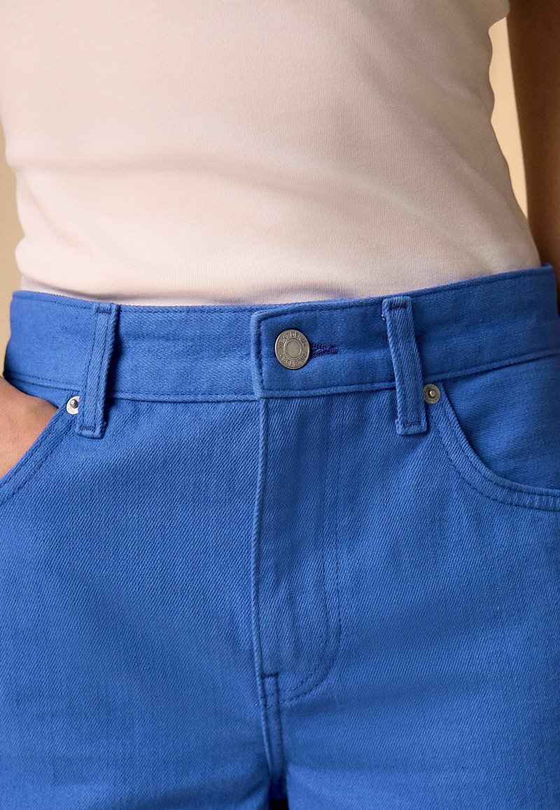 Gros plan d'une personne portant un pantalon bleu vif avec un bouton et des passants de ceinture, associé à un haut beige clair rentré.