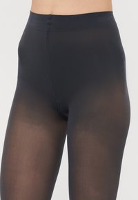 Collants noirs opaques au toucher lisse, à taille haute, avec une ceinture renforcée. Fini translucide près des jambes.