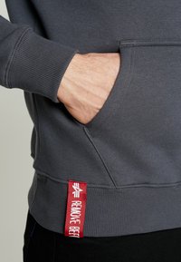 Pilkas džemperis su priekinėmis kišenėmis ir megztu rankogaliu, su ryškiai raudona etikete, kuri skaitoma "PAŠALINTI PRIEŠ SKRYDĮ" šone.