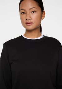 Sweatshirt noir avec un col rond et des manches longues, arborant un logo discret sur la poitrine. Tissu doux avec une texture lisse.