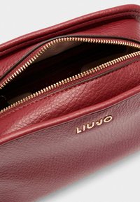 Rode gestructureerde leren tas met gouden ritsen. Voorzien van een logo "LIUJO" dat op de zijkant is ingebrand. Open, waardoor de binnencompartimenten zichtbaar zijn.