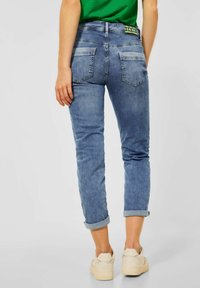 Tmavě modré džíny s přiléhavým střihem, s ohrnutými manžetami a zadními kapsami. Nosené s béžovými teniskami a zelenou košilí.