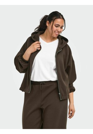 Kvinde smiler, mens hun har en mørkebrun lynlås-hoodie på over en hvid t-shirt og matchende mørkebrune bukser med snøre i taljen.