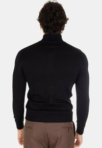 Maglione a collo alto lavorato a maglia nera con texture a costine; design aderente; maniche lunghe; abbinato a pantaloni marroni.