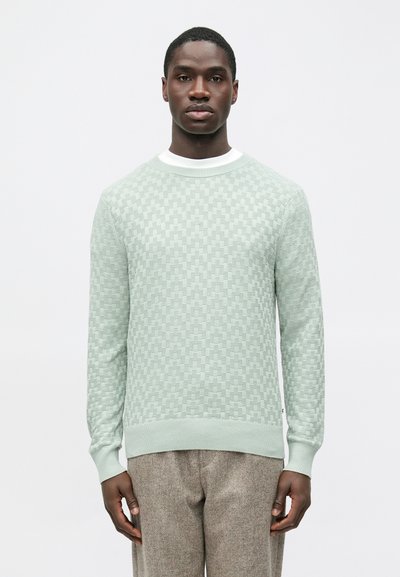 Pull vert clair en tricot avec un motif à carreaux texturé, col rond et poignets côtelés. Associé à un pantalon marron texturé.