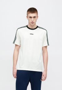 Valge puuvillane T-särk musta ümara kaelusega, millel on varrukatel kolm rohelist triipu ja keskel Adidas logo.