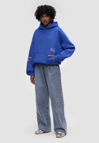 Modrá oversized mikina vyrobená z měkké látky, s přední kapsou a bílými výšivkami. V kombinaci se světle modrými denimovými širokými kalhotami.