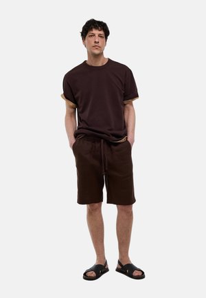 Homme debout, les mains dans les poches, portant un t-shirt marron foncé, un short assorti et des sandales noires sur un fond uni clair.