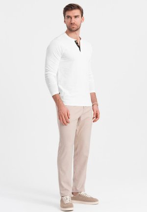 Witte lange mouwen Henley-shirt met zwarte knopenlijst, gecombineerd met lichtbeige broek en beige sneakers, staand tegen een effen achtergrond.