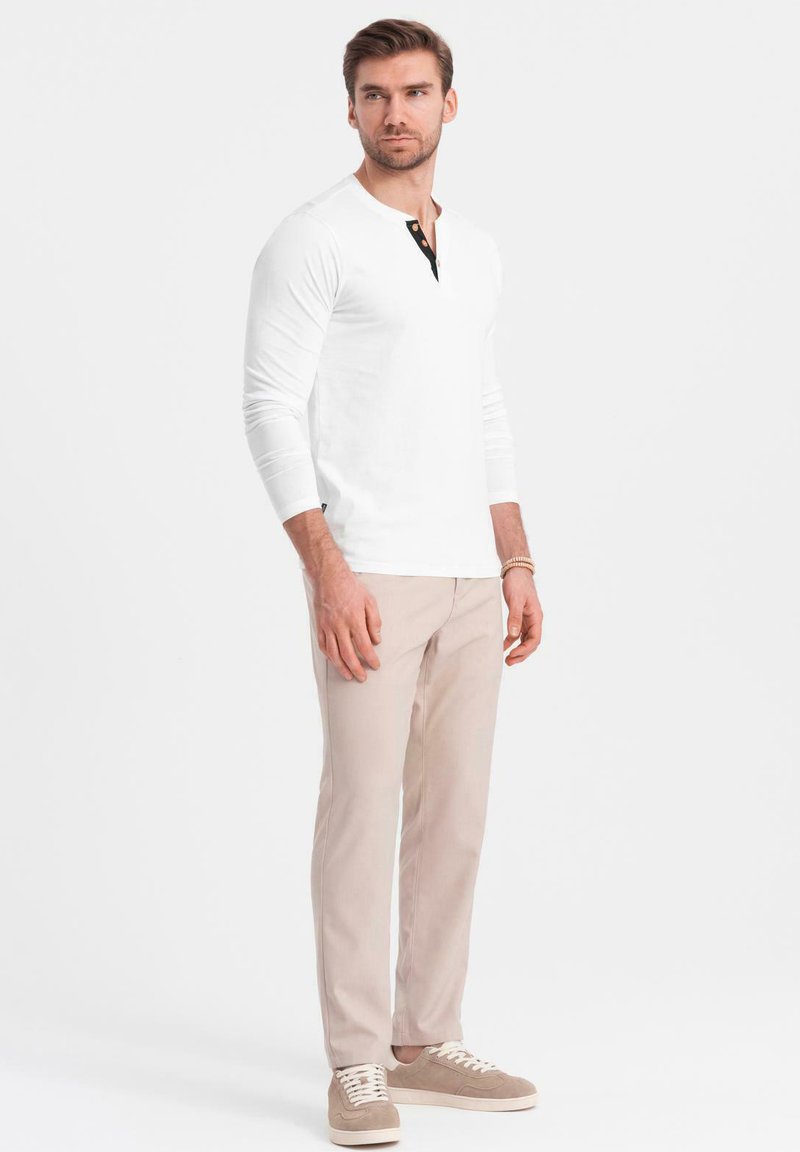 Witte lange mouwen Henley-shirt met zwarte knopenlijst, gecombineerd met lichtbeige broek en beige sneakers, staand tegen een effen achtergrond.