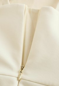 Nærbillede af en elfenbenslynlås, der er delvist åbnet på et tøjstykke af glat stof.