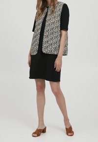 Kobieta w krótkiej czarnej sukience, w kwiecistej bezrękawnik, i brązowych sandałach na obcasie, stojąca na jednolitym białym tle.