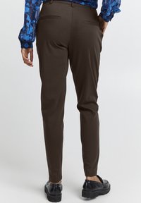 Bruna, slim-fit byxor med en slät yta, två bakfickor, kombinerade med svarta skor. Bakgrunden är neutral och ointrusiv.