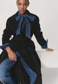 Manteau noir avec des accents en denim et un col à nœud papillon. Manches longues avec des poignets en denim, associé à un pantalon en denim bleu. Texture lisse.