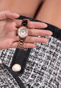 Roosa kuldne randmevaatser metallist rihmaga ja ümmarguse näoga, millel on valge kellanägu, hõbedased aktsendid ja kristallist äär. Käsitsi kuvatud.