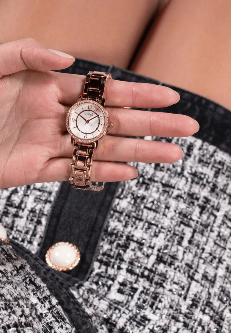 Roosa kuldne randmevaatser metallist rihmaga ja ümmarguse näoga, millel on valge kellanägu, hõbedased aktsendid ja kristallist äär. Käsitsi kuvatud.