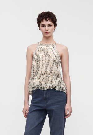 Jeune personne aux cheveux courts et bouclés portant un haut sans manches à motifs avec un ourlet volanté et un jean bleu foncé taille haute, debout devant un fond blanc.