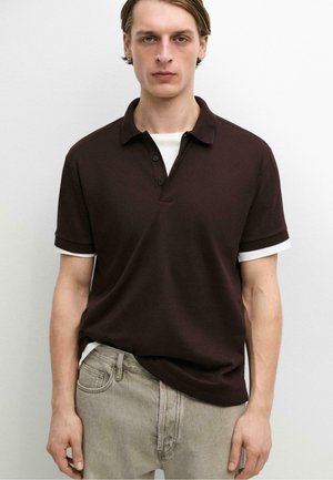 Ung mand iført en mørkebrun kortærmet polo over en hvid t-shirt og lysegrå jeans, står mod en ensfarvet hvid baggrund.