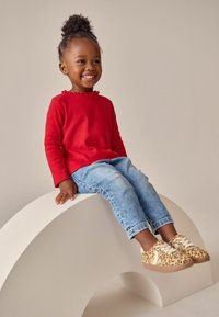 Rød langærmet skjorte med flæsekrave, blå denimjeans og beige leopardmønstret sneakers, siddende på en lysefarvet, buet struktur.