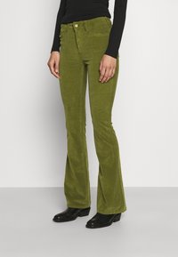 Pantalon en velours côtelé vert évasé avec un bouton doré à la taille et des poches latérales, associés à des bottines noires.