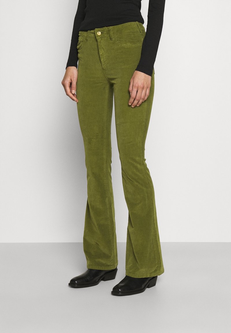 Pantalon en velours côtelé vert évasé avec un bouton doré à la taille et des poches latérales, associés à des bottines noires.