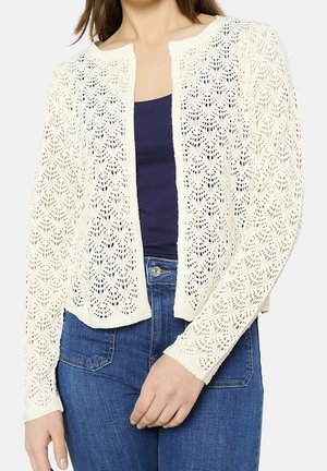 Donna che indossa un cardigan bianco in pizzo aperto sul davanti sopra una maglia blu scuro con jeans blu, in piedi con una mano vicino alla vita.