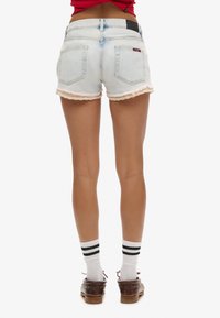 Spijkershort in lichtgewaschte denim met rafelige zoom en kanten rand, voorzien van een zwart patch aan de achterkant, gezien van achteren met witte sokken en schoenen.