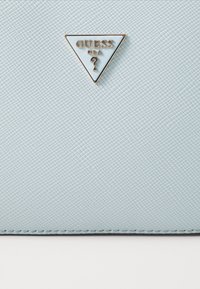 Light blue textured handbag featuring a triangular gold badge with "GUESS USA" and a question mark emblem. Smooth material, sleek design.