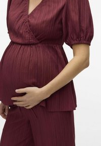 Blusa da maternità in borgogna con texture, maniche a palloncino corte, scollatura a V e vestibilità morbida, abbinata a pantaloni coordinati. La mano poggia sul pancione.