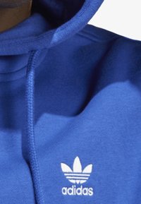 Sininen huppari, joka on valmistettu pehmeästä kankaasta. Hupparin rintakehässä on valkoinen Adidas-logo ja kolme raitaa.