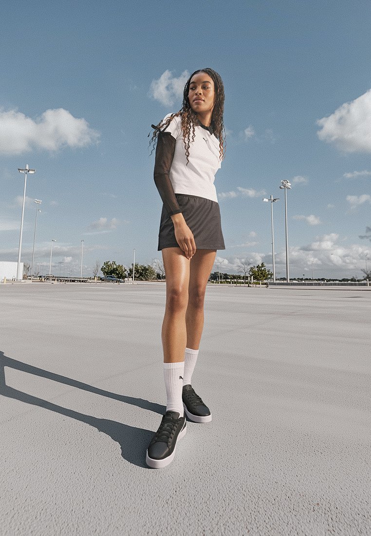 Des chaussures de sport noires, des chaussettes blanches jusqu'aux chevilles, un haut blanc avec des manches noires et une jupe noire. Le mannequin se tient sur une surface plane sous un ciel bleu.