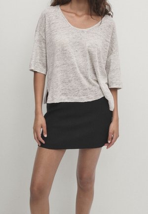 Femme portant un haut ample gris clair à manches courtes, légèrement rentré dans une mini-jupe noire ajustée, sur un fond uni.