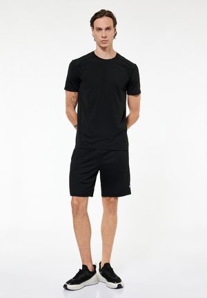 Mężczyzna stoi z rękami za plecami, ubrany w czarną sportową koszulkę, czarne spodenki i czarno-białe buty sportowe na białym tle.