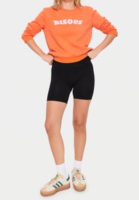 Orange sweatshirt med vit "BISOU"-tryck, matchad med svarta cykelshorts. Modellen bär krämfärgade sneakers med gröna detaljer och vita strumpor.