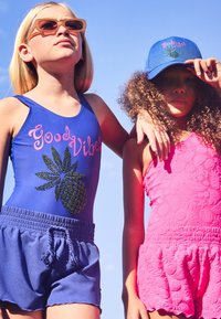 Du vaikai maudymosi kostiumėliuose: vienas dėvi mėlyną vientisą kostiumą su ananaso dizainu ir užrašu "Good Vibes", o kitas - rožinę tekstūruotą romperį.
